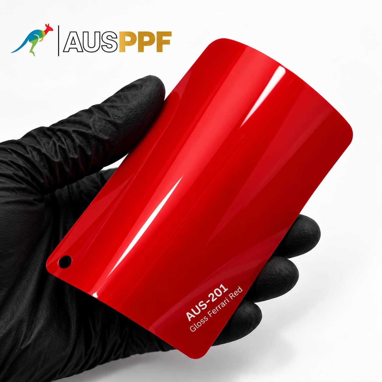 A gloved hand holding a swatch of AusPPF AUS-201 Gloss Ferrari Red paint protection film, showing its deep red gloss finish.