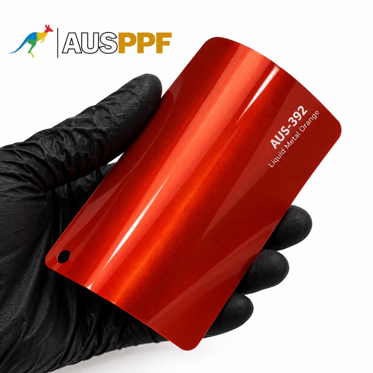 A gloved hand holding a swatch of AusPPF AUS-392 Liquid Metal Orange paint protection film, showing its metallic orange finish.